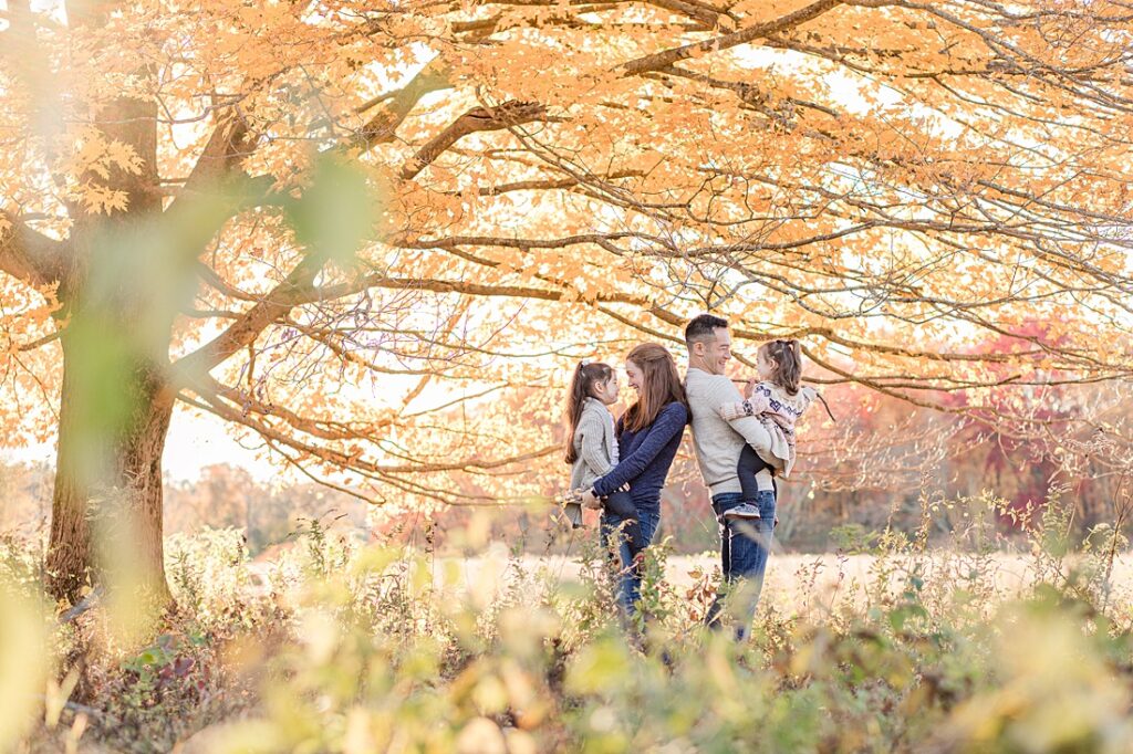 Parents stand back to back holding children under tree with fall foliage during family photo session in Wayland Massachusetts