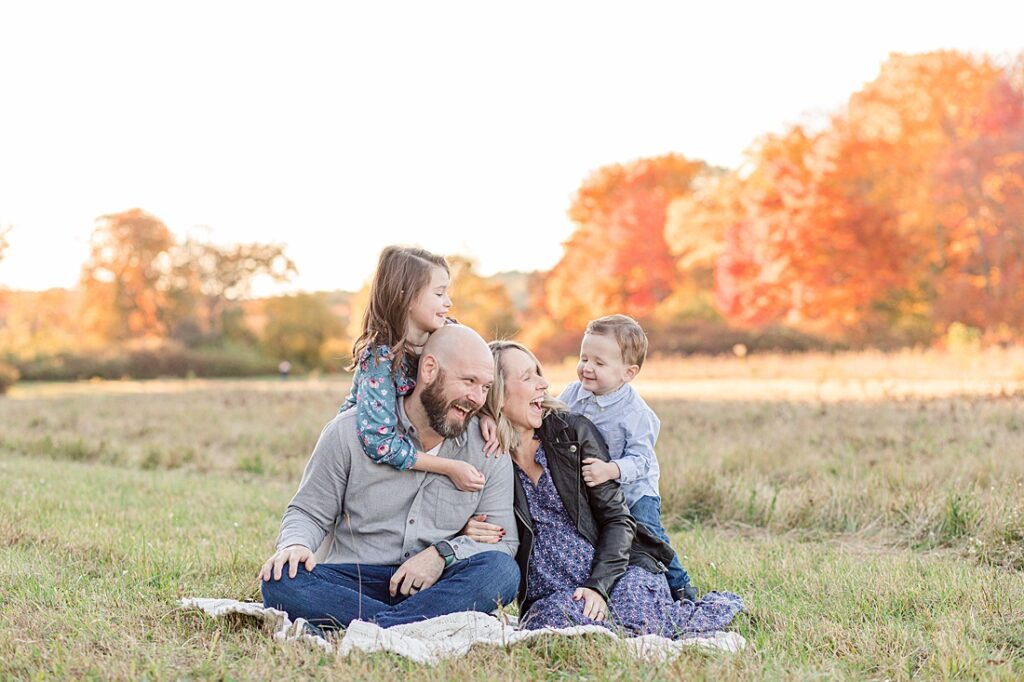 Family laughs together sitting on a blanket in a field in front of fall foliage in Wayland Massachusetts