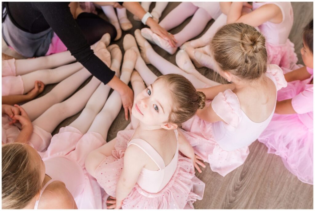 Ballerina's sit in circle with feet together, one girl looks up at camera at DanceFIT Natick 