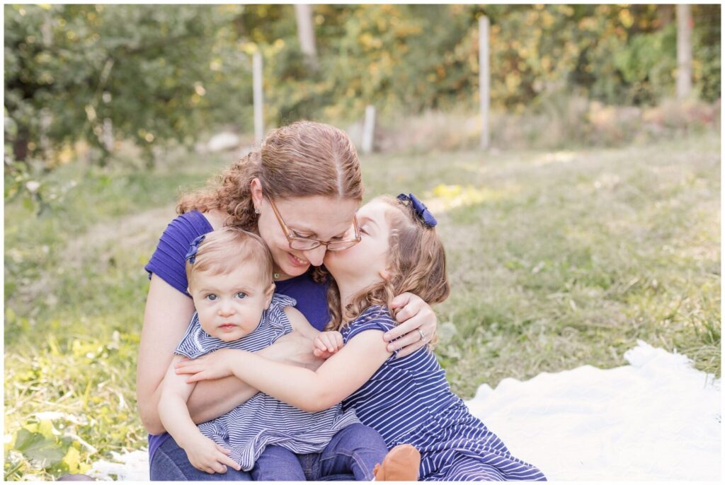 Mom cuddles on blanket in orchard with daughters at Dowse Orchard Sherborn MA during fall family photo session