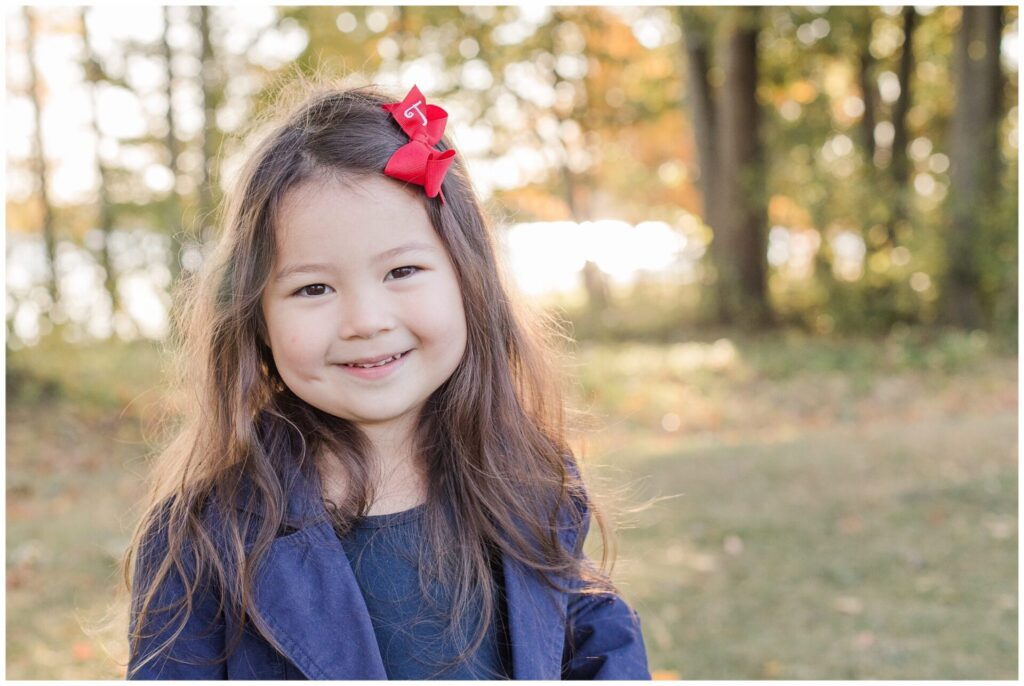 girls with red bow smiles at camera during fall family photo session. morning light reflects of water behind her at Farm Pond Park Framingham MA