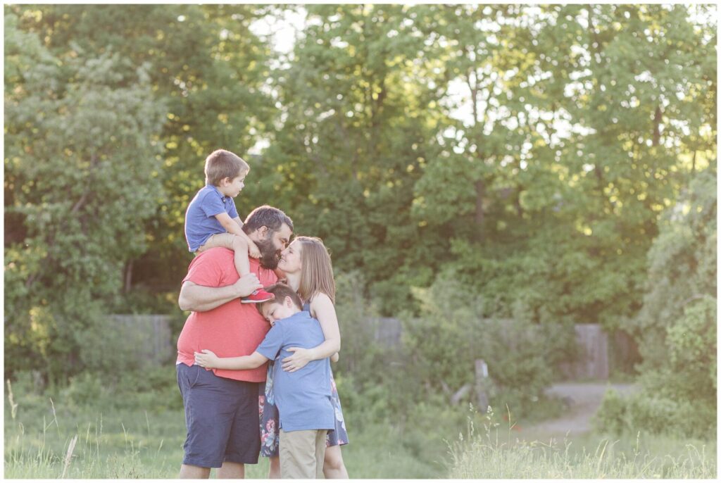 One boy sits on dad shoulders while other hugs parents as they kiss during family photos in Needham MA