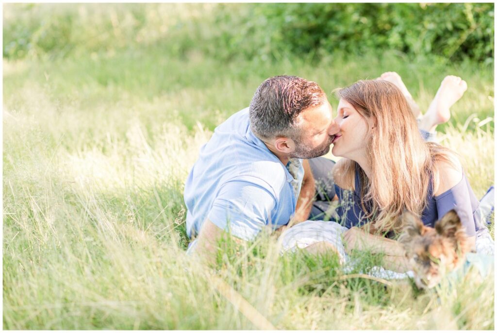 couple laying in grass kisses over baby with dog next to them for newborn photos 