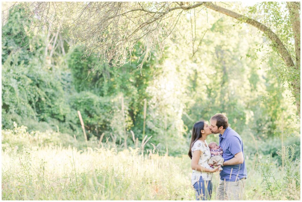 parents kiss while holding newborn during outside newborn photo session in Massachusetts