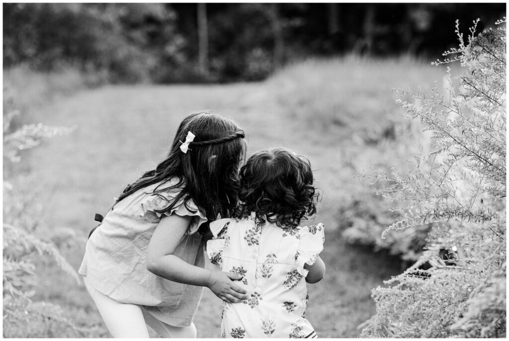 Lack and white photos of sisters walking through field in Newton Massachusetts. Big sister hand on little sister back. 
