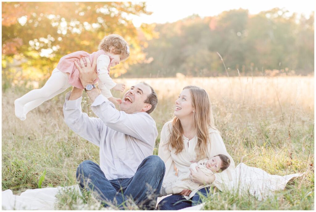 family sits on blanket in field and dad holds toddler up in the air for family photo in Wayland Massachusetts
