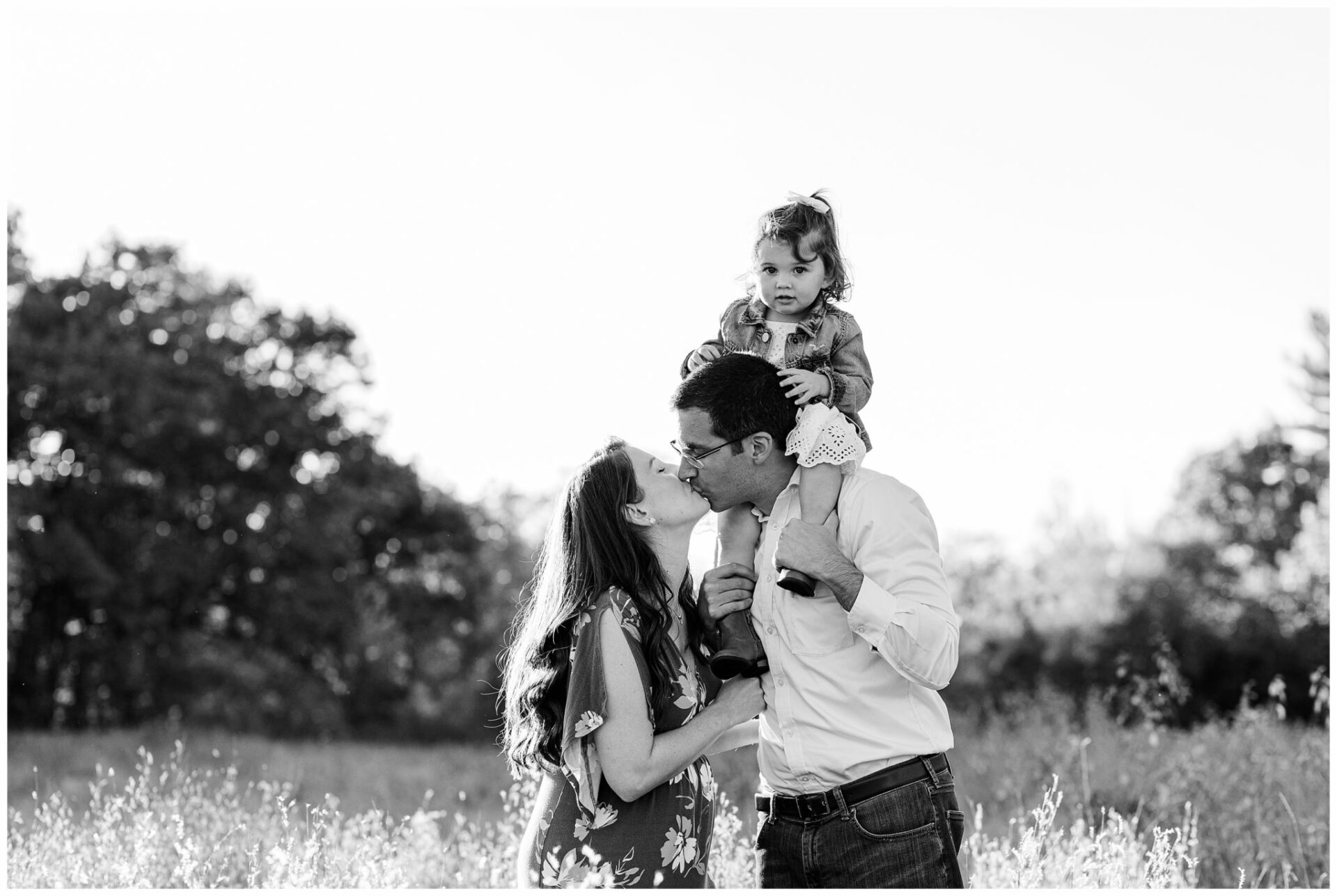 black and white photo of dad with daughter on shoulders kissing pregnant wife in field in Wayland Massachusetts.