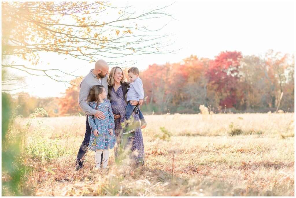 Natick Massachusetts family laughs together in field with fall foliage during maternity photo session. 