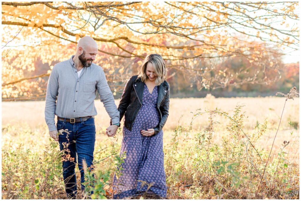 Natick Massachusetts couple holds hands walking while soon to be mom of three holds pregnant belly for maternity photo session. 