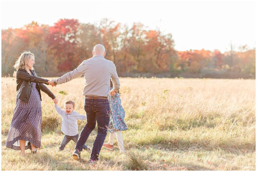 Soon to be mom of three holds hand with family walking in a circle in a field for family photos in Wayland Massachusetts