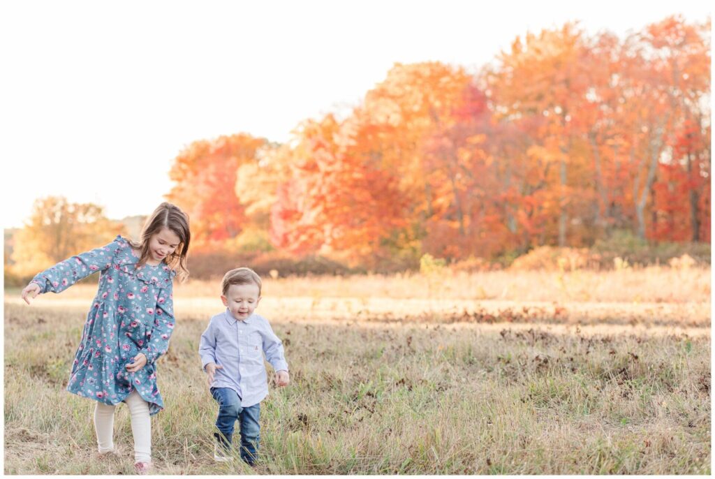 Brother and sister walk together though field with foliage behind them for family photo session in Wayland Massachusetts. 
