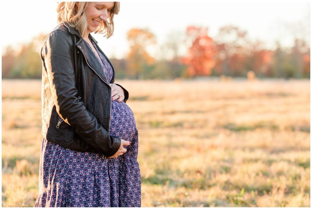 Natick soon to be mom of three stands in field holding pregnant belly for maternity photo session. 