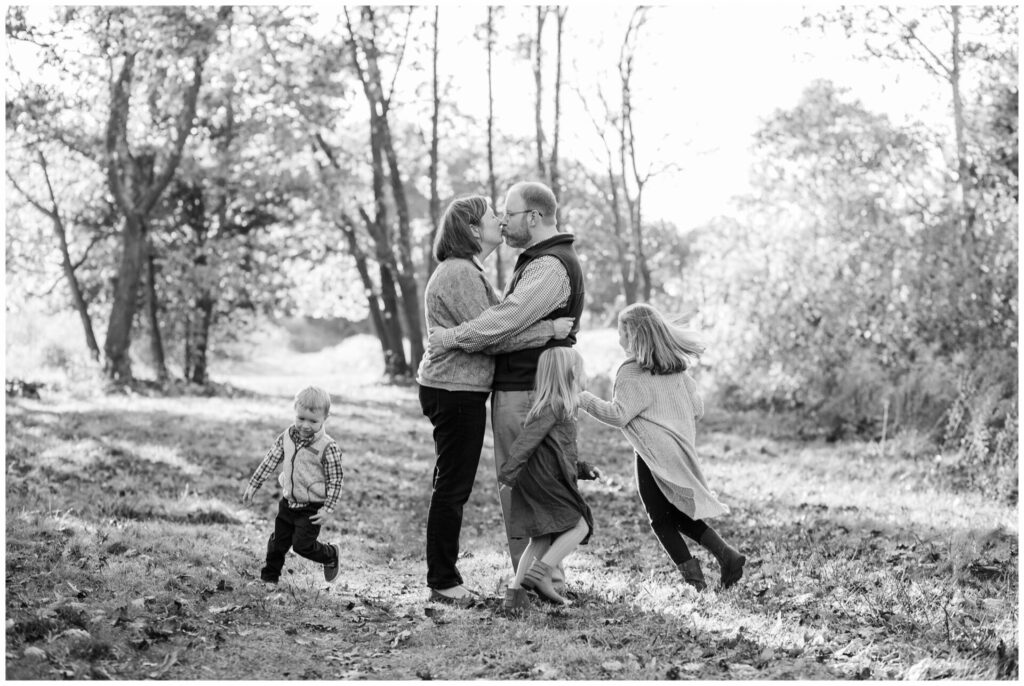 Black and white photo prompt of parents kissing while kids run around them during Natick Massachusetts family photo session