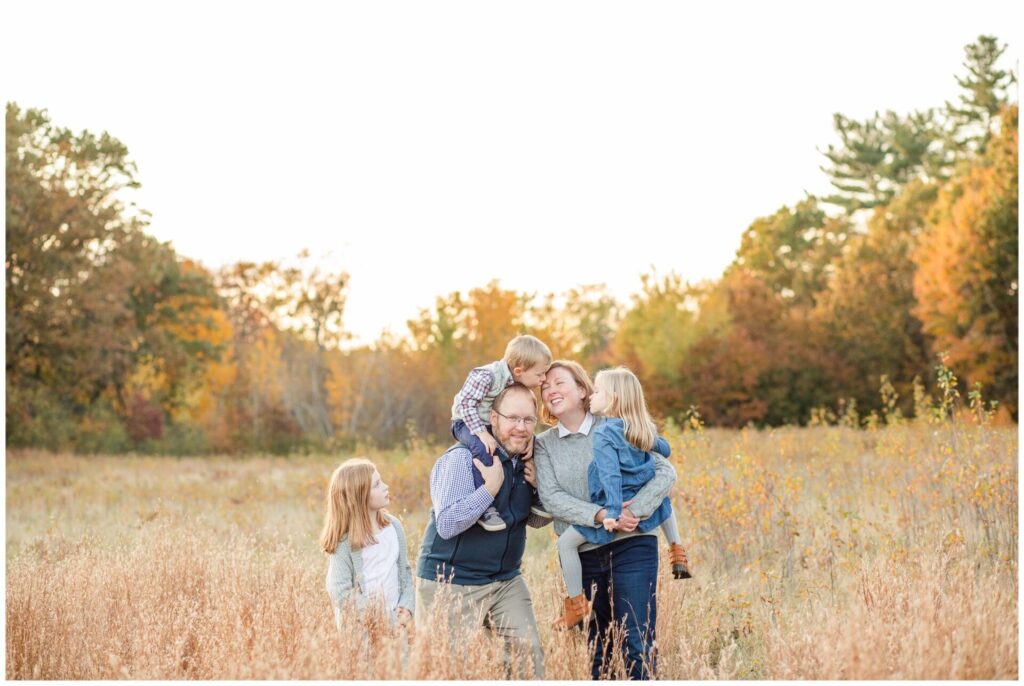 Natick Massachusetts family photo in field son on dads shoulders kisses mom while she holds daughter