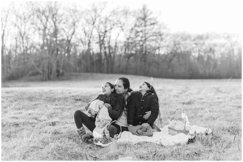 black and white photo of mother and daughters sitting on blanket looking up at clouds at Wayland MA Conservation Land. 