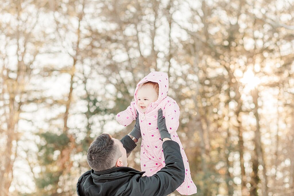 Dad holds baby in snow suit up in the air with sun in background during one year old birthday photos at Norfolk Airfield, Norfolk Massachusetts