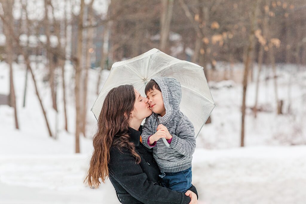 Mom kisses son holding umbrella in the snow during family photo session in Natick Massachusetts for NICU blog