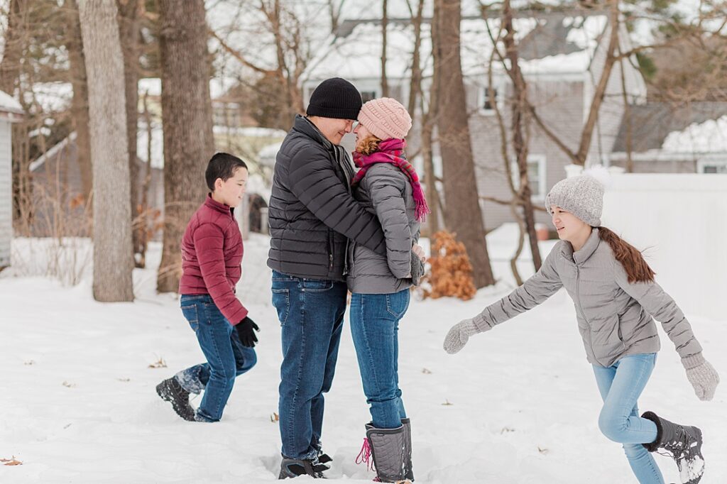Parents kiss while kids run around them in the snow during family photo session for NICU blog in Natick Massachusetts