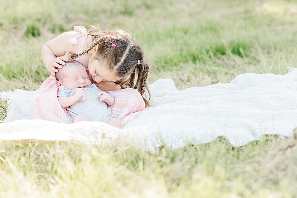 Girl kisses newborn baby brother while sitting on a blanket during newborn photo session with Sara Sniderman Photography at Barber Reservation, Sherborn Massachusetts