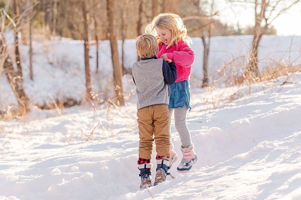 siblings hold hands and jump in snow for family photographer at Wayside Inn Grist Mill, Sudbury MA