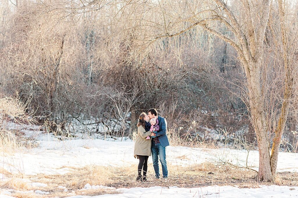 Parents kiss while holding daughter in snowy field for family photographer NICU experience blog at Barber Reservation, Sherborn MA