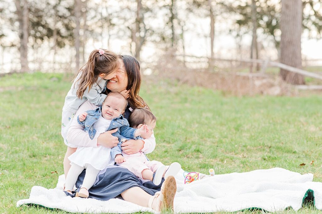 Mom holds twin babies while toddler daughter kisses her face during family photo session with Sara Sniderman Photography at Barber Reservation, Sherborn Massachusetts