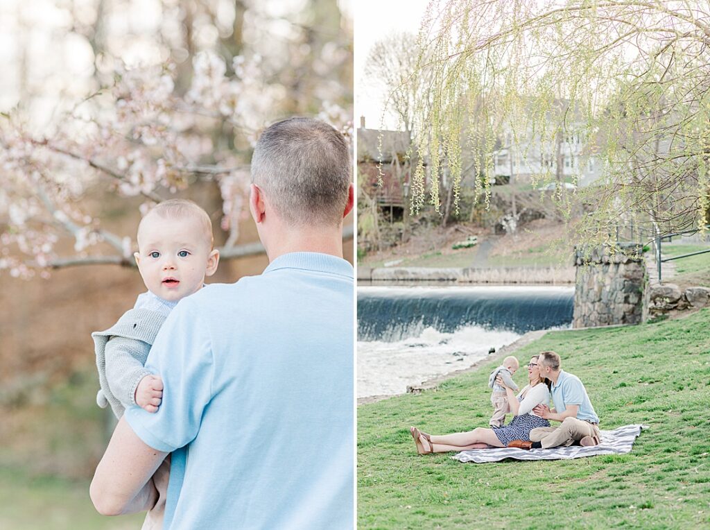 Father holds baby over shoulder in front of flowering tree and family sits on blanket together during family photo session with Sara Sniderman Photography at South Natick Falls, Natick Massachusetts