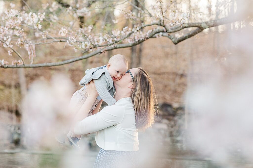 Mom kisses baby in front of blooming tree with flowers in the foreground during family photo session with Sara Sniderman Photography for NICU blog at South Natick Falls, Natick Massachusetts