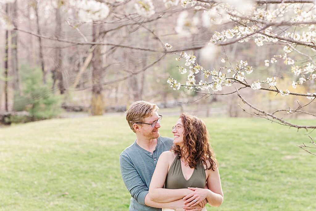 couple smile at each other under blooming tree during family photo session with Sara Sniderman Photography in Sherborn Massachusetts