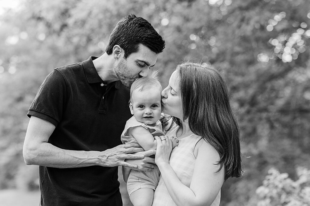 black and white photo of parents kissing babies head during photo session at Elm Bank, Wellesley Massachusetts with Sara Sniderman Photography