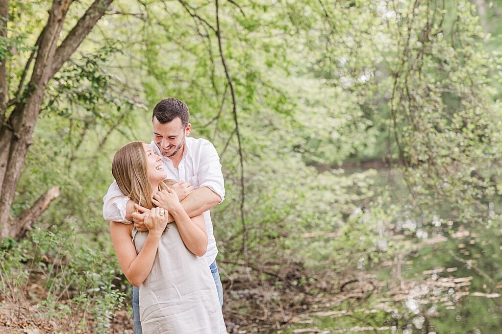 couple smiles at each other while standing in front of water during photo session at Elm Bank, Wellesley Massachusetts with Sara Sniderman Photography