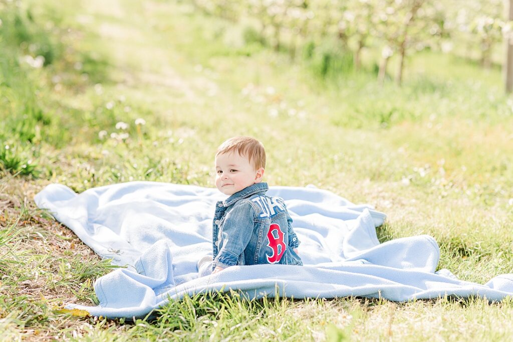 Baby wearing Boston Red Socks jacket smiles over his shoulder while sitting in orchard during family photo session with Sara Sniderman Photography at Dowse Orchard, Sherborn Massachusetts
