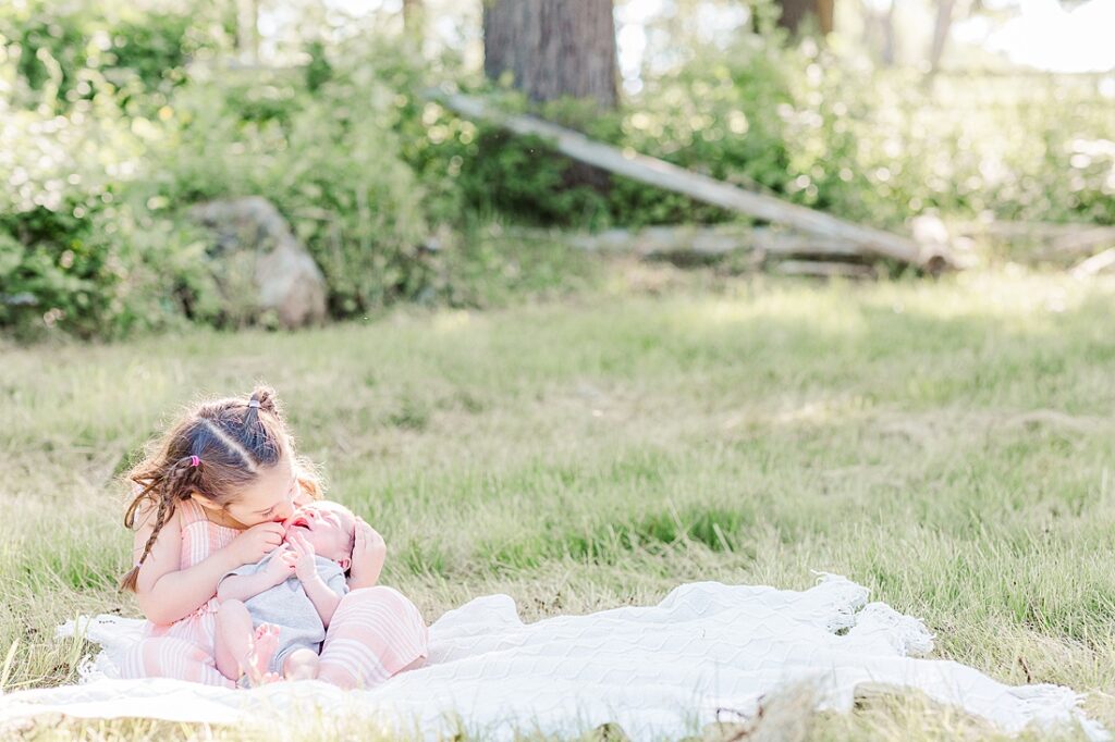 sibling photo of big sister holds newborn brother sitting on a blanket in a field during outdoor newborn photo session with Sara Sniderman Photography at Barber Reservation, Sherborn Massachusetts. 