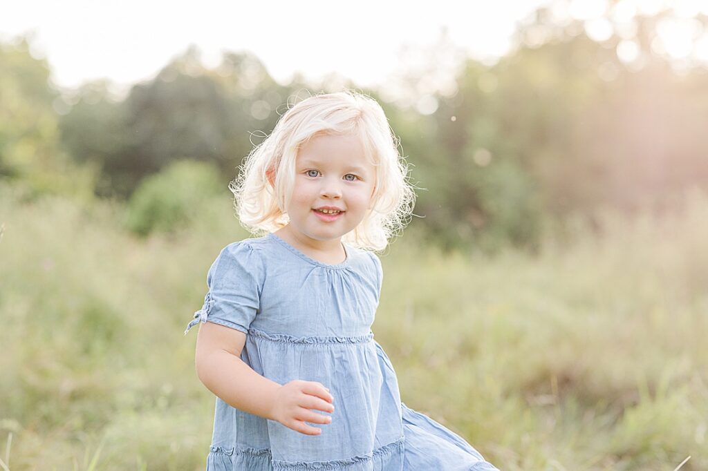 Toddler girl smiles at camera while running through field at golden hour during extended family photo session with Sara Sniderman Photography at Oak Grove Park, Millis Massachusetts