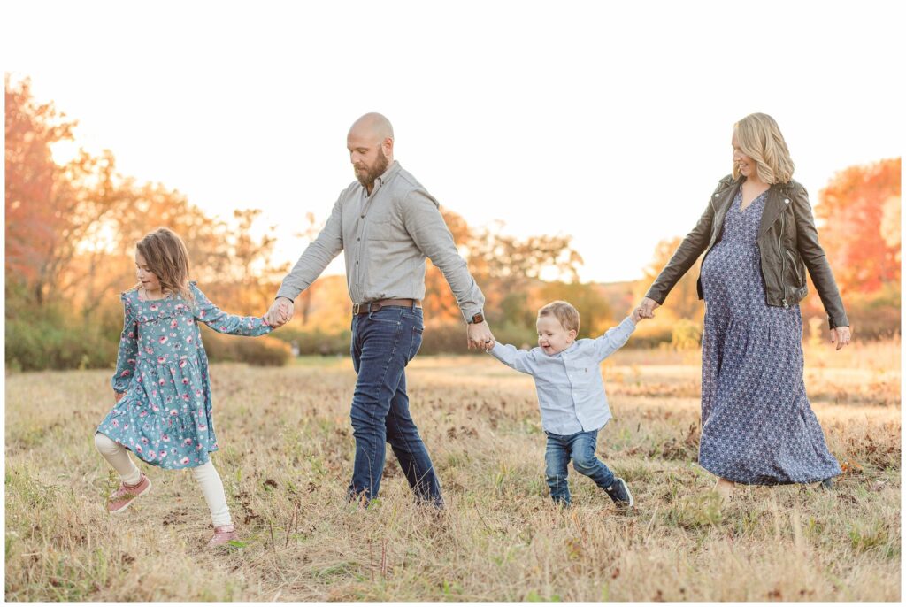 family walks hand in hand across field during maternity photo session with Sara Sniderman Photography in Natick Massachusetts