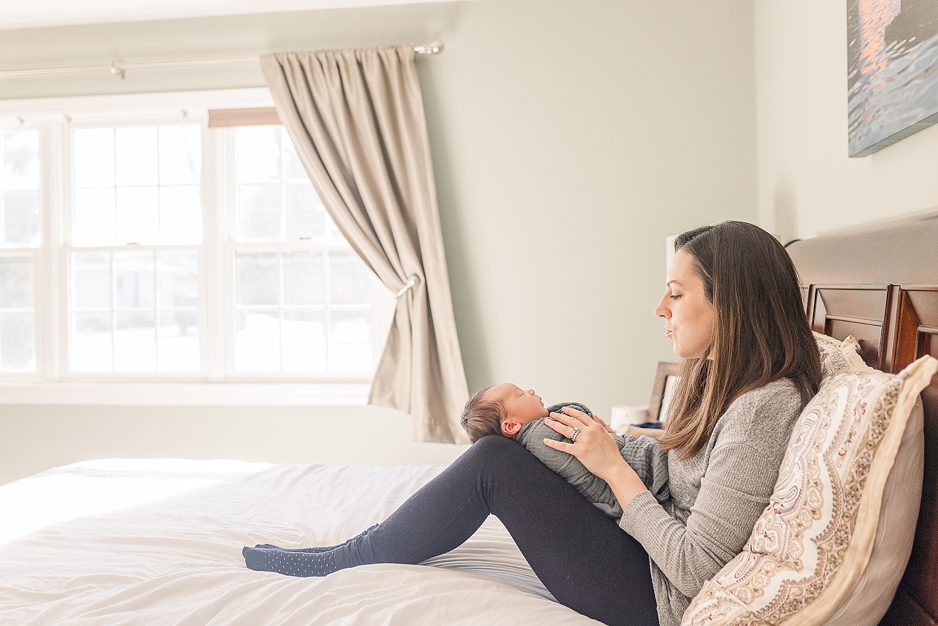Mom sits on bed with baby during in home newborn photo session with Sara Sniderman Photography in Framingham Massachusetts