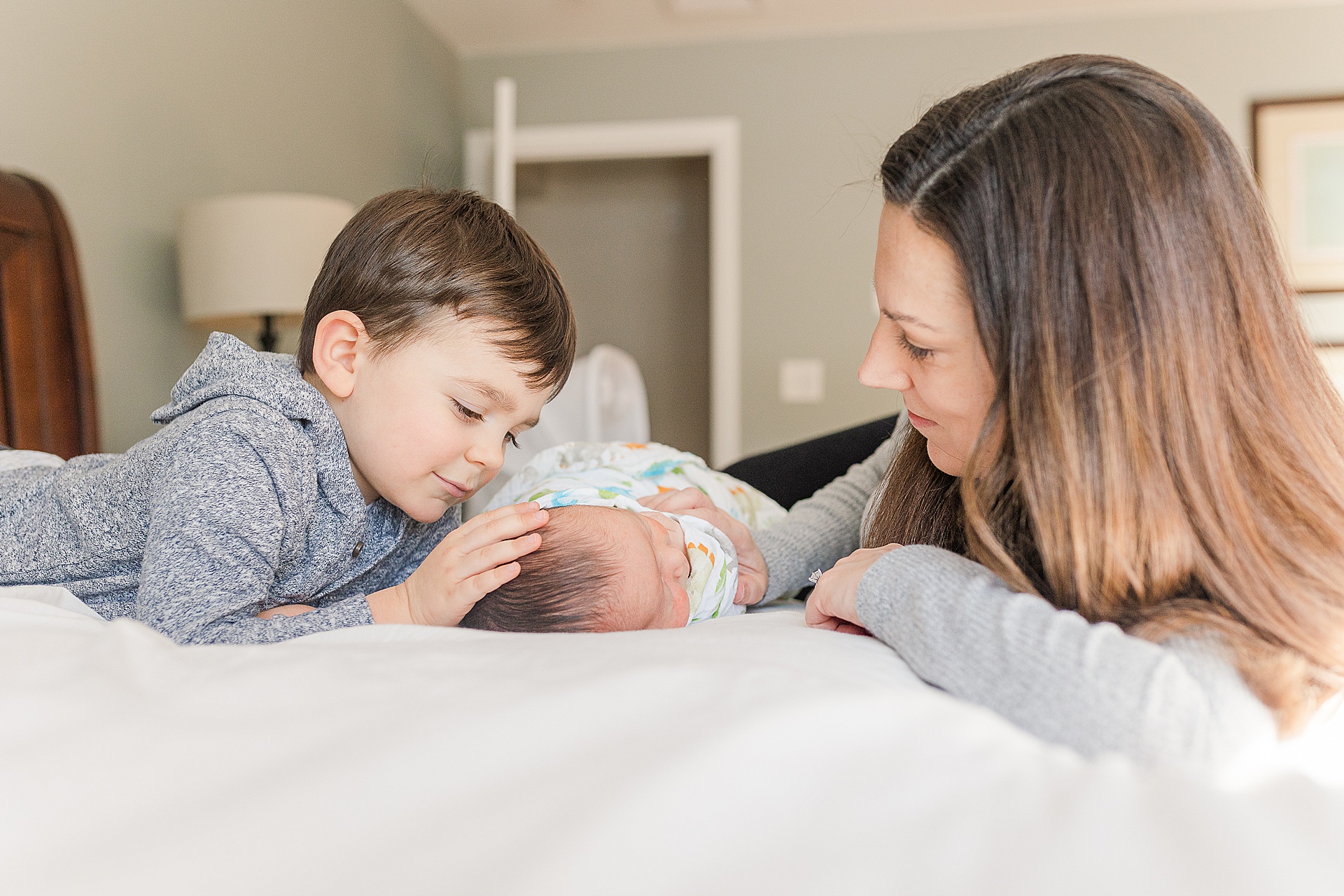 Mom and big brother lay on bed with baby during in home newborn photo session with Sara Sniderman Photography in Framingham Massachusetts