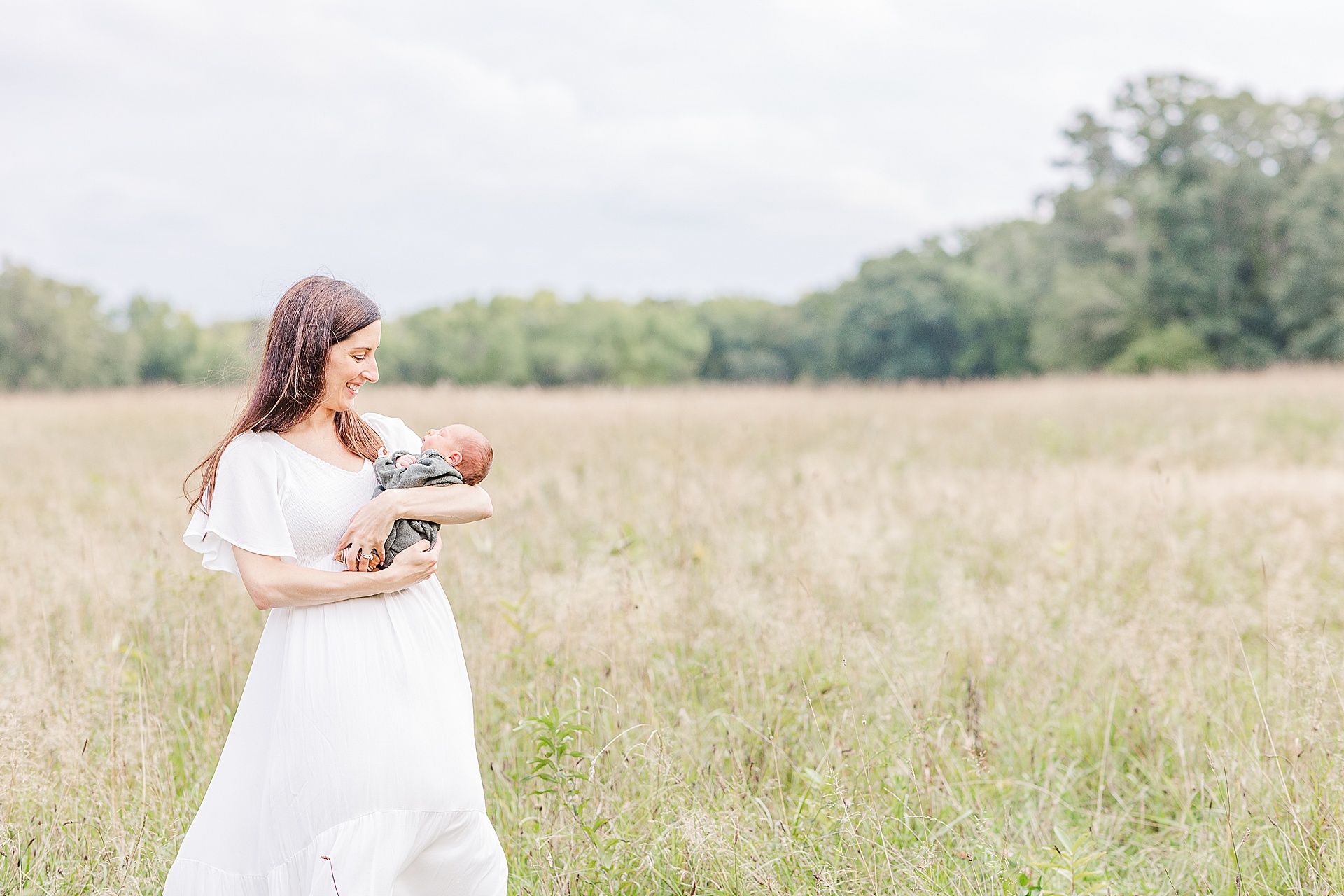 Mom stands in field smiling down at newborn baby during outdoor newborn photo session with Sara Sniderman Photography at Heard Farm, Wayland Massachusetts
