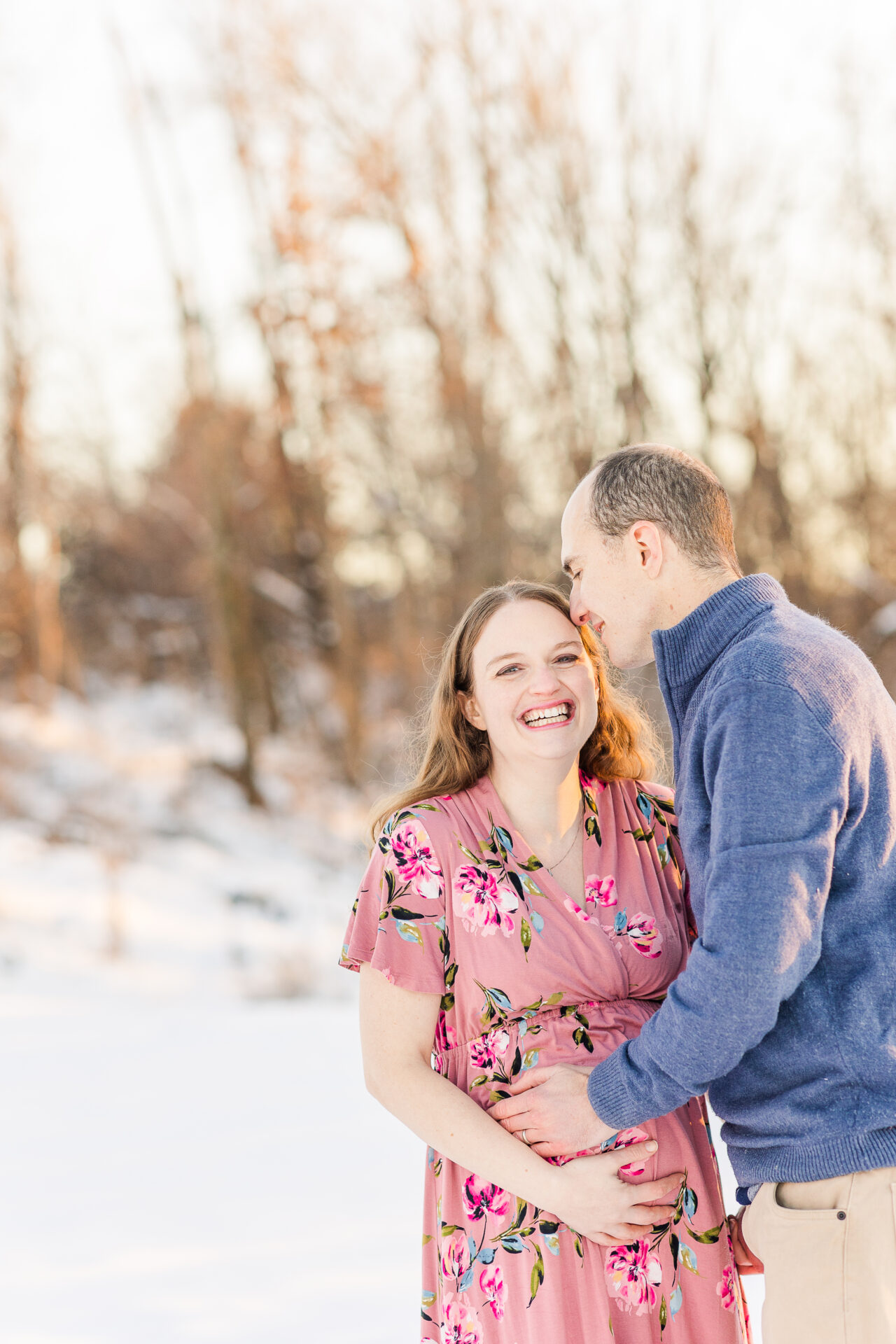 pregnants woman laughs while husband kisses her head during snowy maternity session with Sara Sniderman Photography at Medfield State Hospital in Medfield Massachusetts