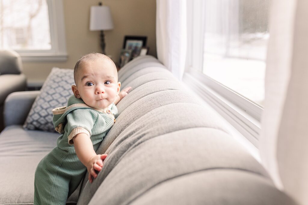 baby stands on couch looking out window during in home photo session with Sara Sniderman Photography in Natick Massachusetts