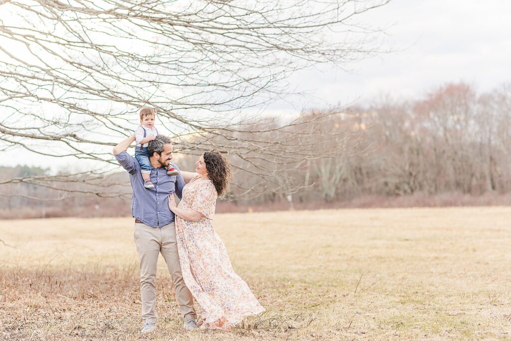 toddler sits on dad shoulders and parents smile at each other during family maternity session for pregnancy announcement in Natick Massachusetts with Sara Sniderman Photography