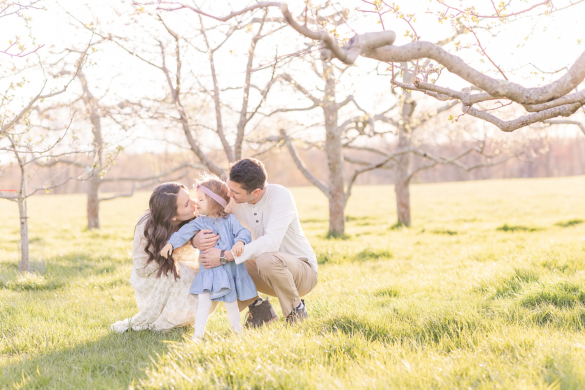 parents snuggle daughter during apple blossom family photo session at Heard Farm in Wayland Massachusetts with Sara Sniderman Photography