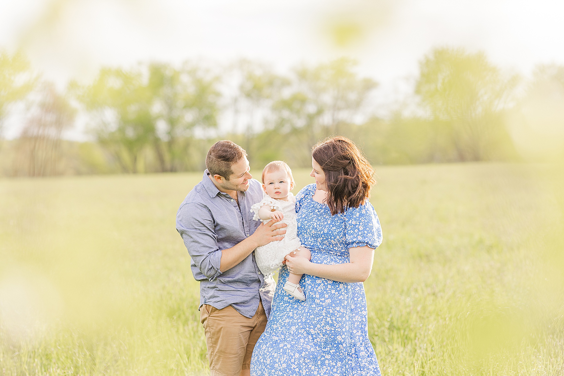 Parents smiles at one year old in field during first birthday family photo session with Sara Sniderman Photography at Heard Farm in Wayland Massachusetts