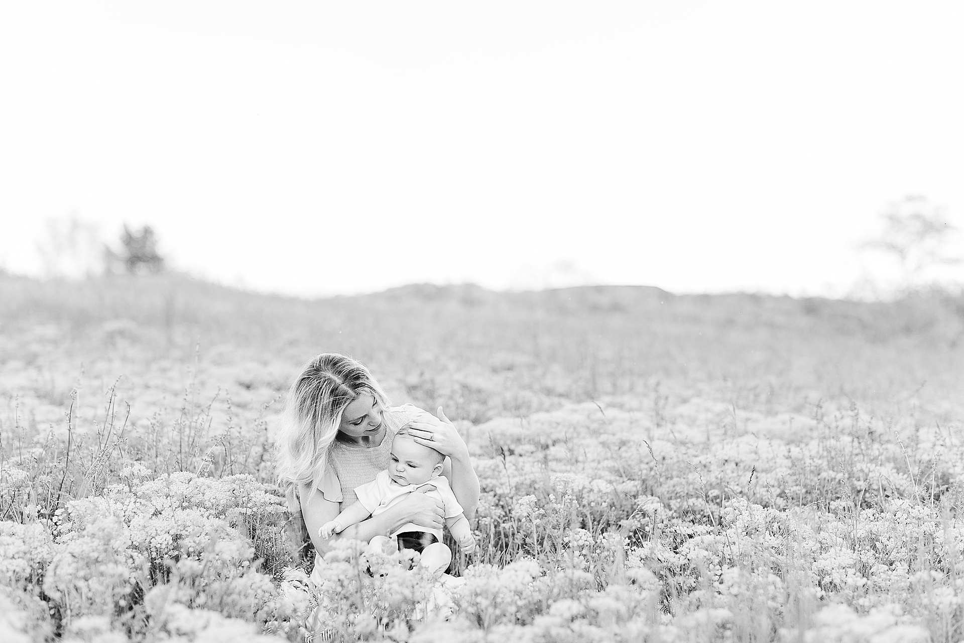 black and white photo of mother sitting in field of flowers with her baby during 6 month old photo session with Sara Sniderman Photography at Charles River Peninsula in Needham Massachusetts