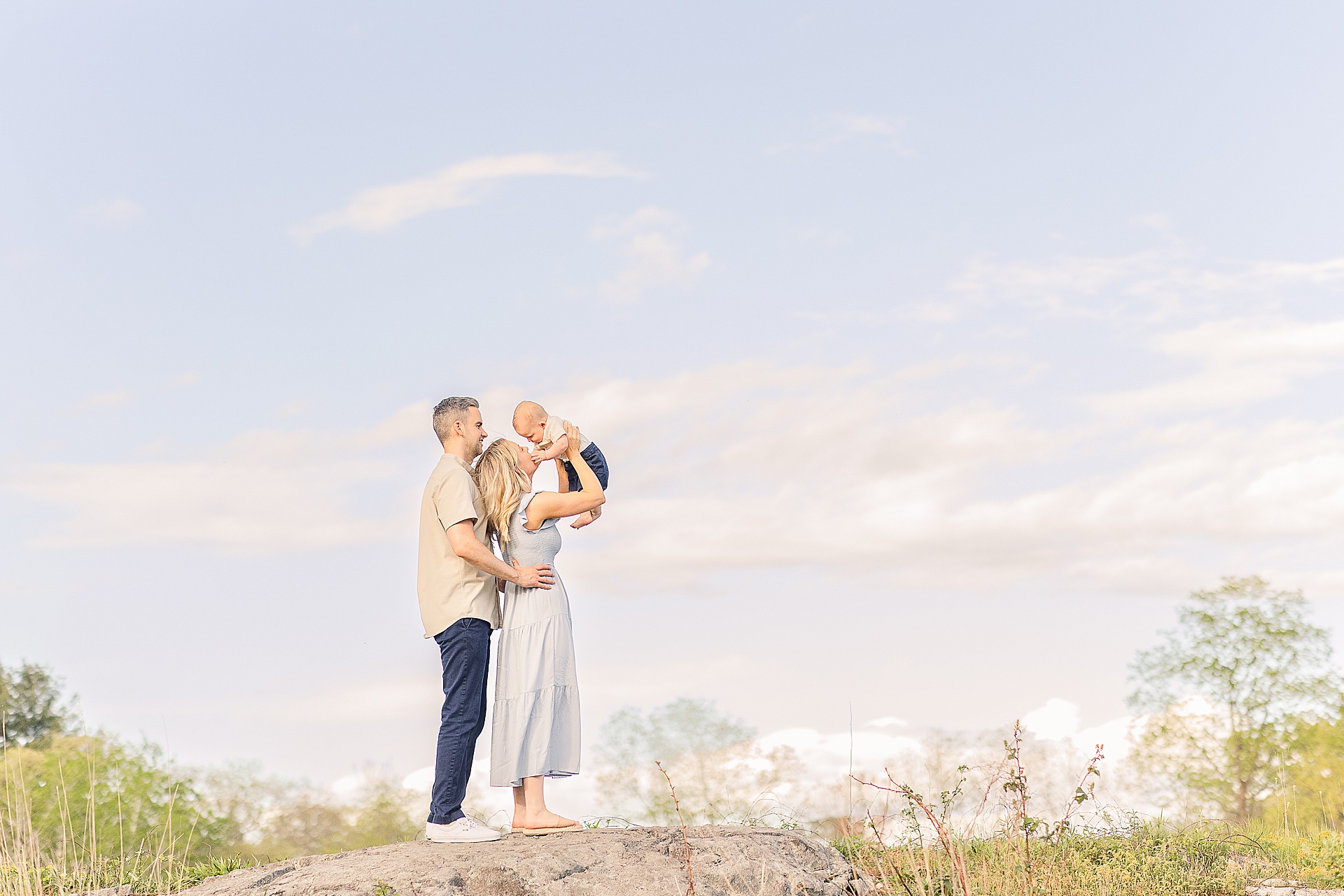parents look up at baby while standing on a rock during 6 month old photo session with Sara Sniderman Photography at Charles River Peninsula in Needham Massachusetts