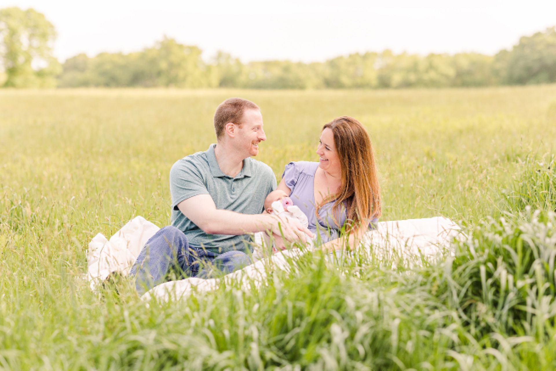 Parents cuddle with newborn on a blanket in a field of grass during outdoor newborn photo session with Sara Sniderman Photography at Heard Farm Wayland Massachusetts