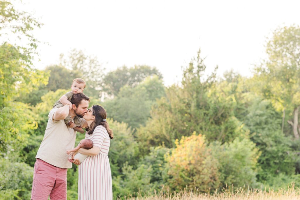 dad holds toddler on shoulders and kisses wife who is holding baby during outdoor newborn photo session with Sara Sniderman Photography at Oak Grove Park Millis Massachusetts