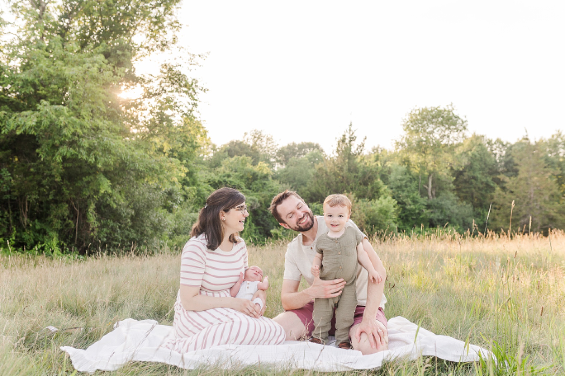 family sits in field together during outdoor newborn photo session with Sara Sniderman Photography at Oak Grove Park Millis Massachusetts