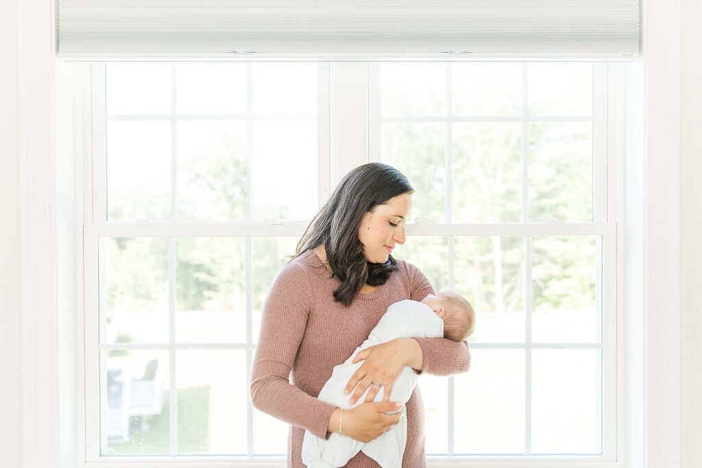 mother holds baby in front of window during in home newborn photo session with Sara Sniderman Photography Southborough Massachusetts