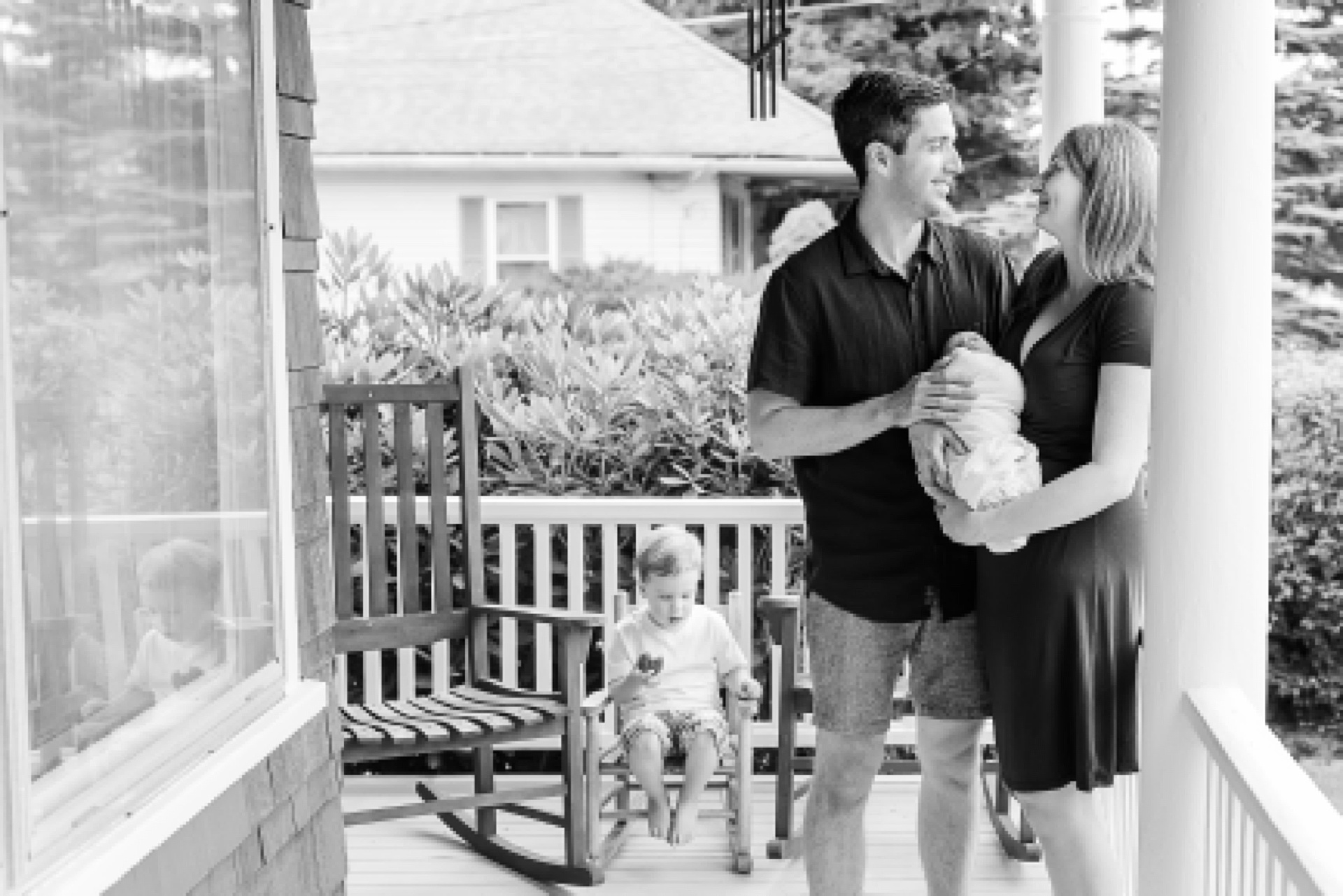 black and white of parents holding baby on porch while toddler eats cup cake behind them during in home newborn photo session with Sara Sniderman Photography in Ashland Massachusetts